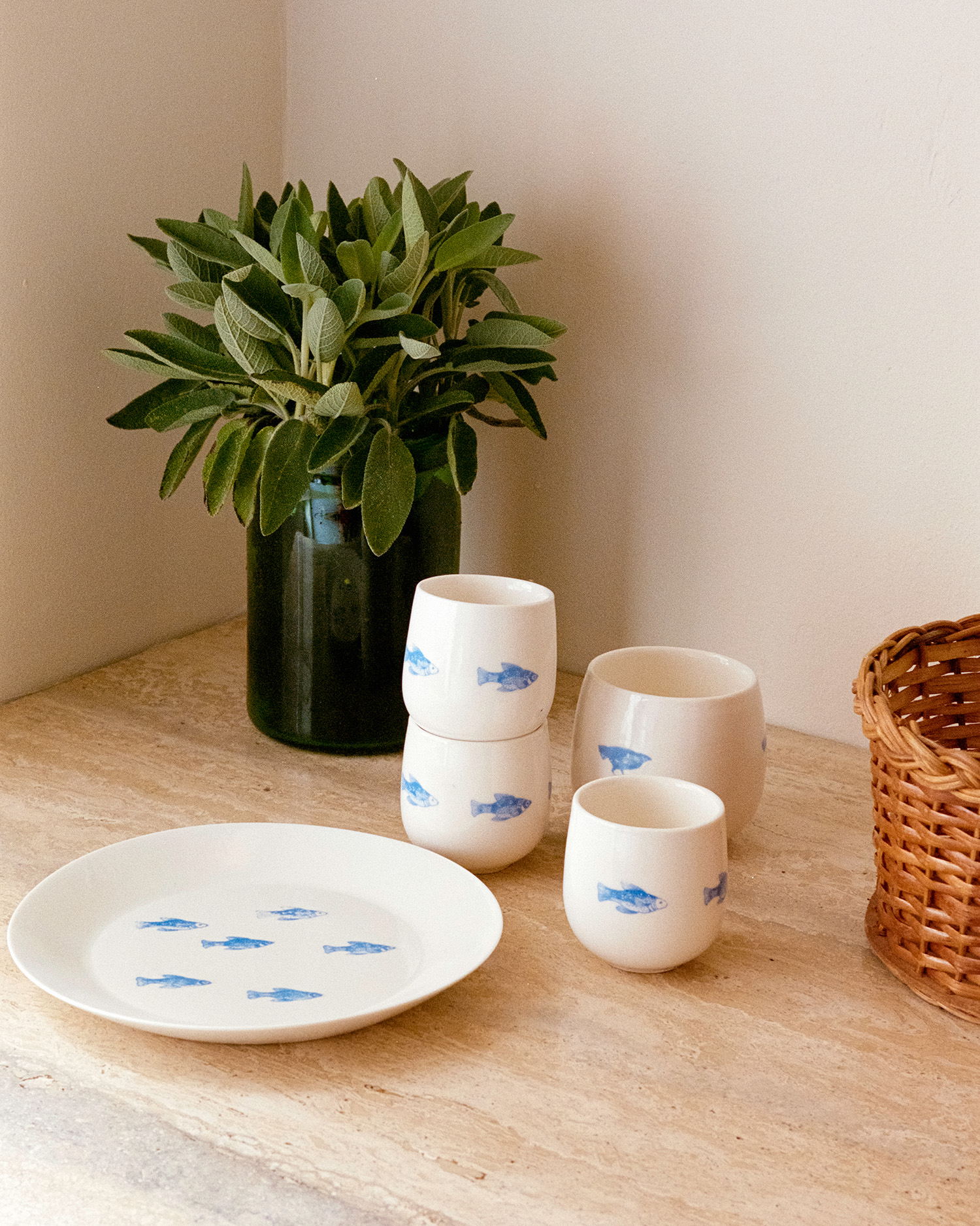 Hand-painted ceramic cups and plate with blue fish motifs beside a green vase with sage on a travertine counter