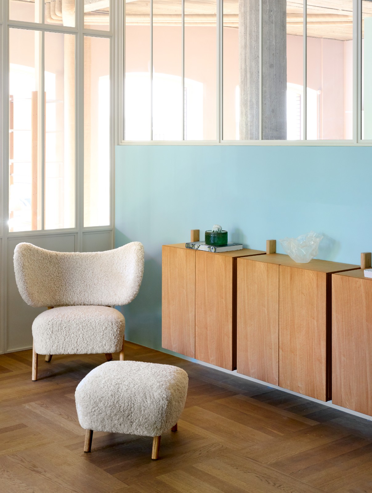TMBO Lounge Chair with pouf in white sheepskin beside oak cabinets against a light blue wall