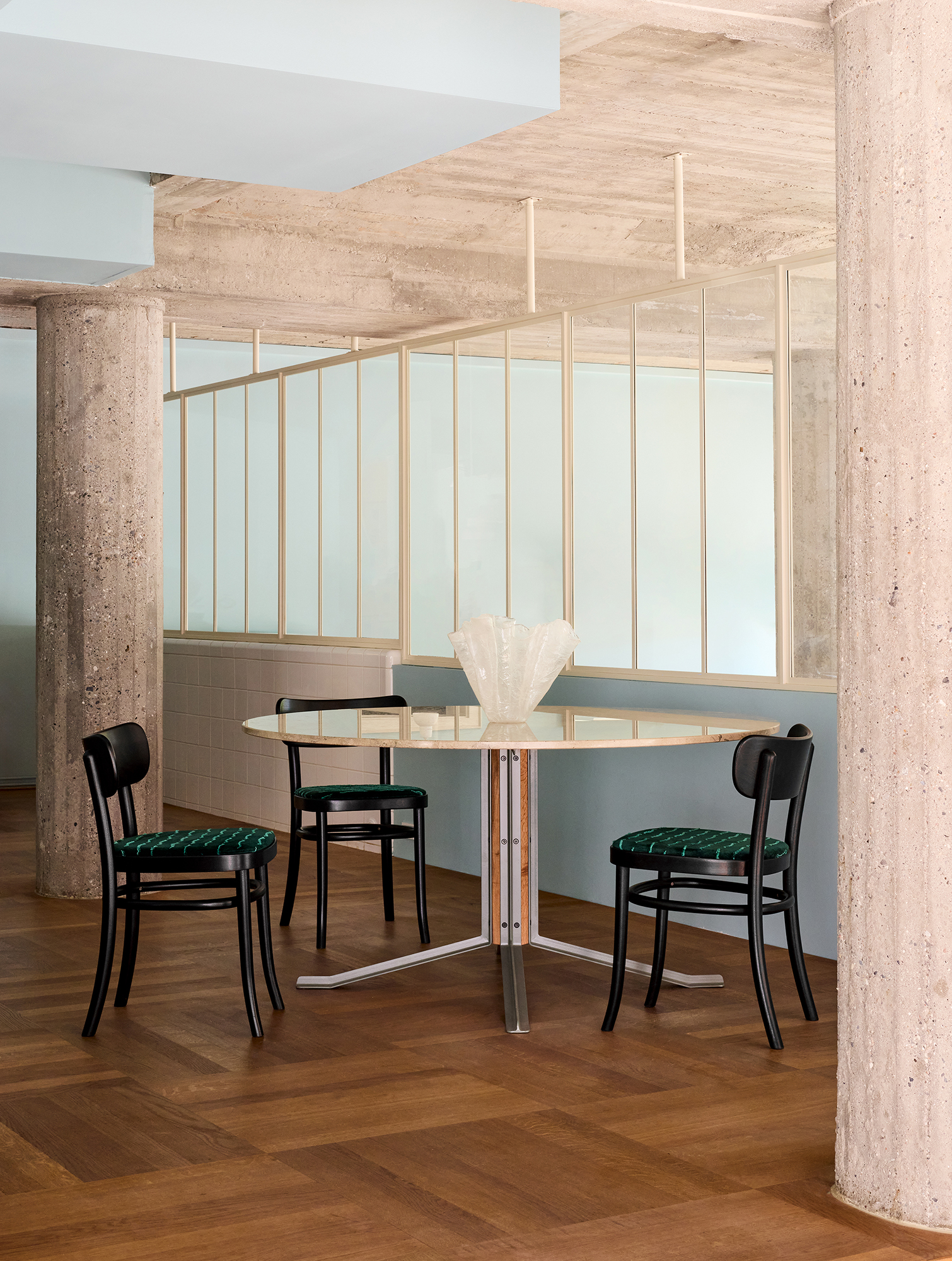 Black MZO Chairs with green velvet seat pads around a round travertine table in a concrete room