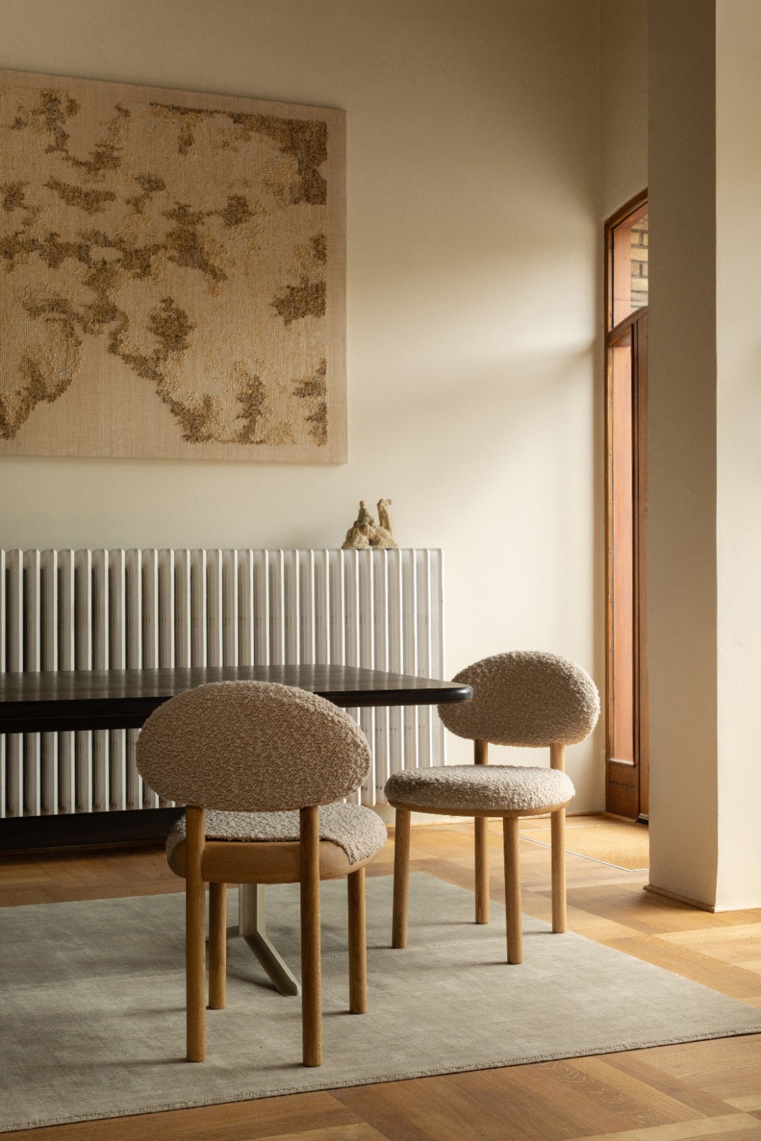 Two MAG Dining Chairs in cream bouclé at a dark dining table beneath a large tapestry