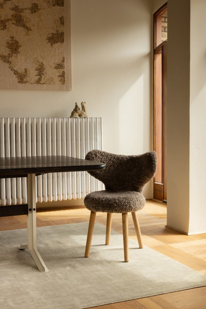 WNG Chair in brown sheepskin at a dark dining table in front of a radiator and tapestry