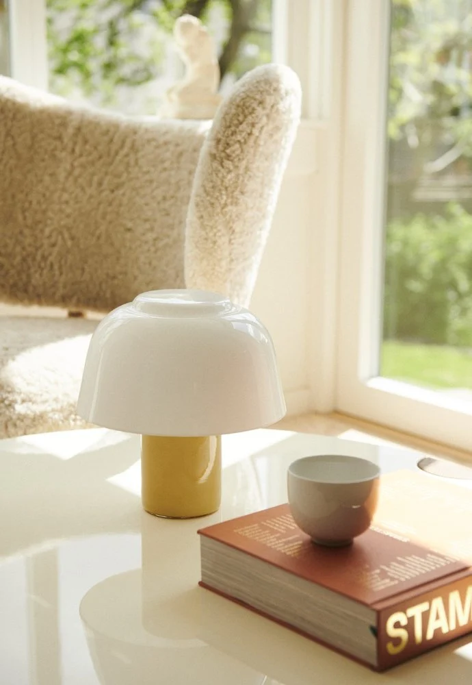 YUKI Lamp Olive on a round coffee table beside a book and ceramic cup by a sunlit window
